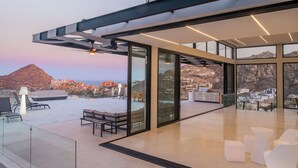 A modern rooftop terrace with glass walls, offering panoramic views of a mountainous landscape and a coastal town. - Luxury Villa w/ Tennis, Pickleball, Pool, Hot Tub & Fitness Room, Views (Cabo San Lucas)