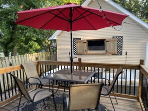 A deck with a table and chairs, a red umbrella, and a house with shutters in the background.