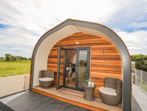 Une structure moderne, partiellement ouverte, en bois, dotée d’une porte en verre, de deux fauteuils en osier et d’une petite table sur laquelle est posée une plante.