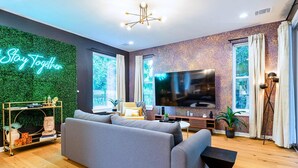 Una sala de estar moderna con un sofá gris, un televisor de pantalla plana, una pared verde con un cartel que dice 'Stay Together' y ventanas amplias.