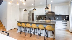 Una cocina moderna con una isla central, taburetes de bar, lámparas colgantes y electrodomésticos de acero inoxidable.