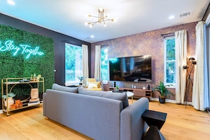 A modern living room with a grey sofa, a flat-screen TV, a green wall with a 'Stay Together' sign, and large windows.