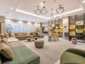 Un vestíbulo moderno con un sofá modular, una mesa de centro y una zona de estar con sillas. Hay lámparas colgantes geométricas y una pared con estantes que contienen plantas y libros.