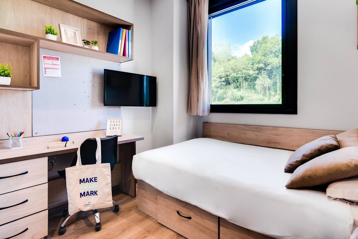 A bedroom with a bed, a desk with a computer, a chair, and a window with a view of trees.