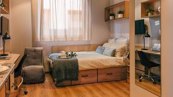 A compact bedroom with a bed, desk, chair, and a small shelf with plants.