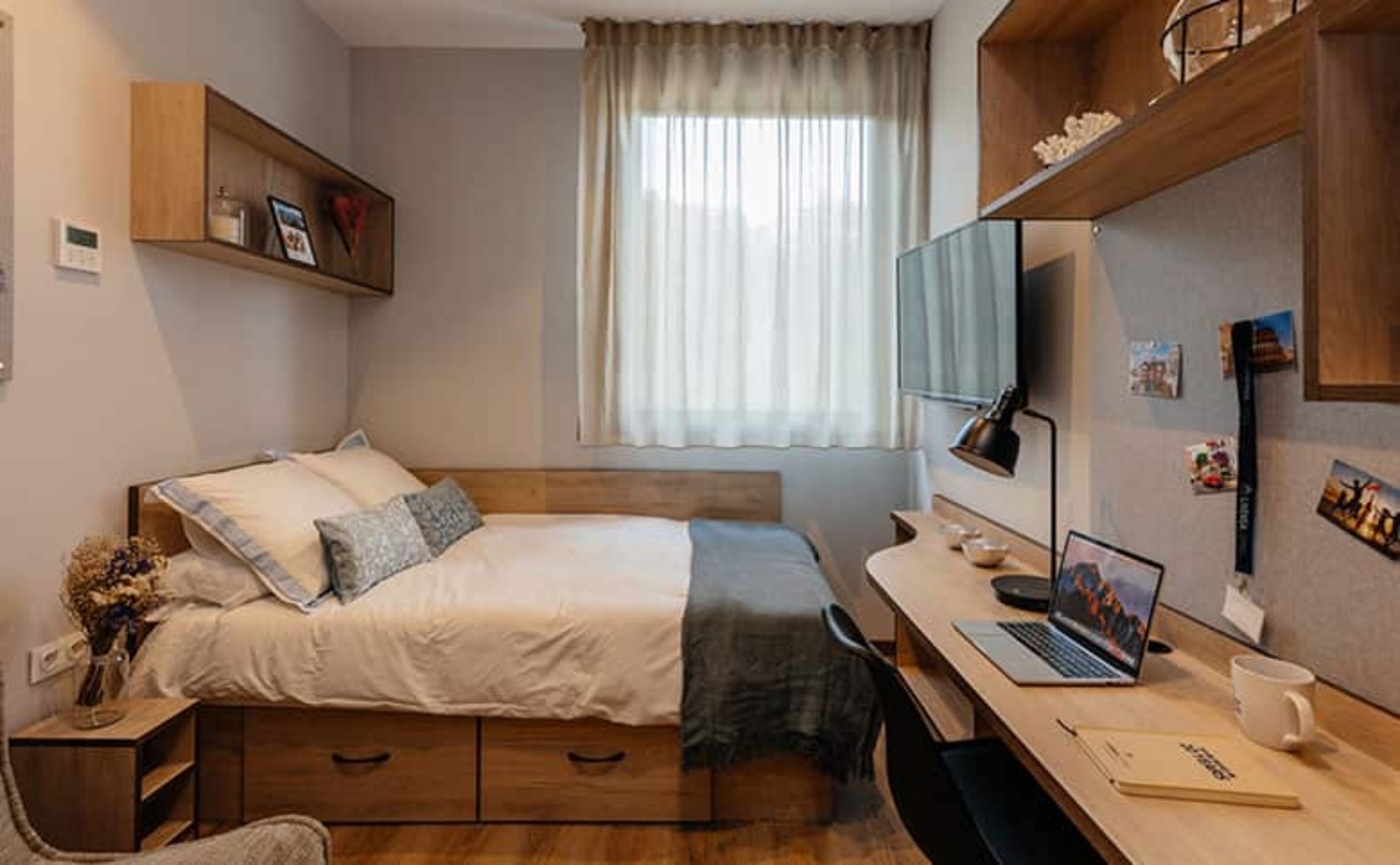 A bedroom with a bed, bedside table, desk, laptop, and a window with curtains.