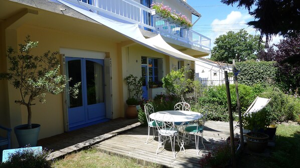 Un edificio amarillo con puerta y ventanas azules, un balcón con plantas en macetas y una terraza de madera con muebles de exterior.