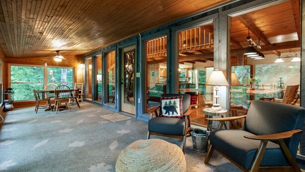 A spacious room with a wooden ceiling, large windows offering a view of the outdoors, a dining area with a table and chairs, and a cozy seating area with a lamp and a chair. - Quietside Cabin: Sunroom, Fire Pit, Theater Den, & Stunning Views (Walland)