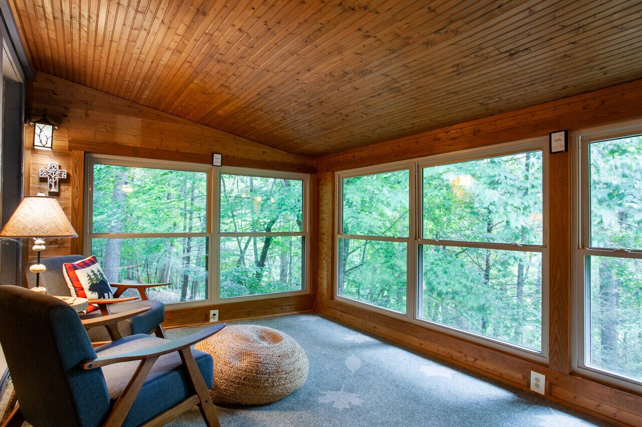 Una camera da chalet accogliente con ampie finestre, soffitto in legno, una poltrona confortevole, un tavolino e una lampada.