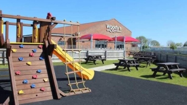 Ein Spielplatz mit Kletterwand und Rutsche, Picknicktischen und einem Gebäude mit dem Schild 'Woody's'.