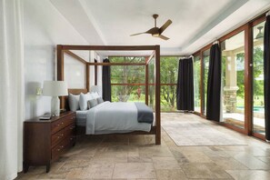 A spacious bedroom with a four-poster bed, a wooden nightstand, and a large window offering a view of the outdoors.
