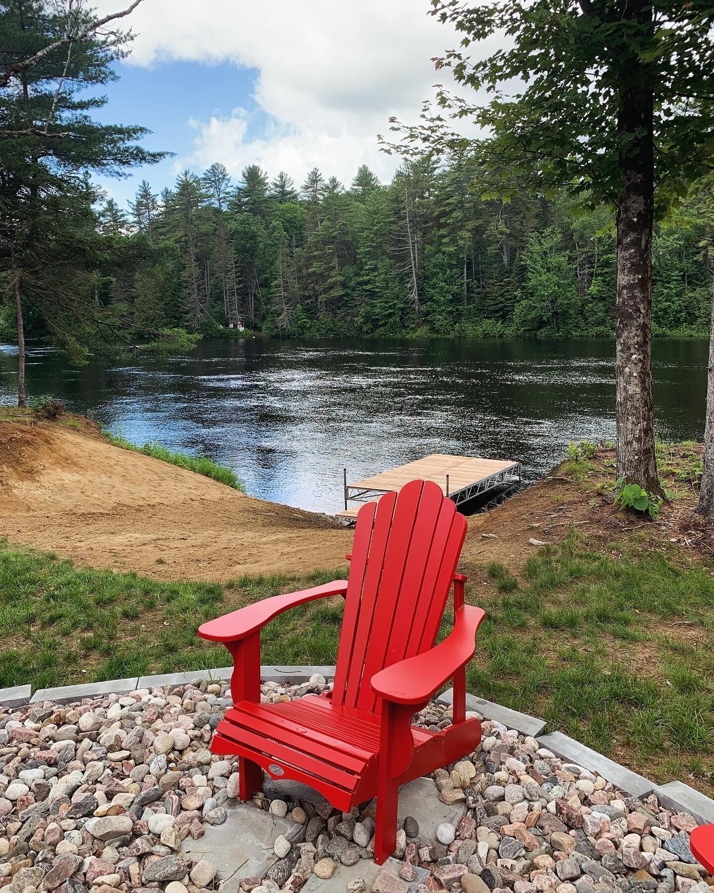 A red Adirondack chair by a lake with a wooden dock and a pile of rocks.