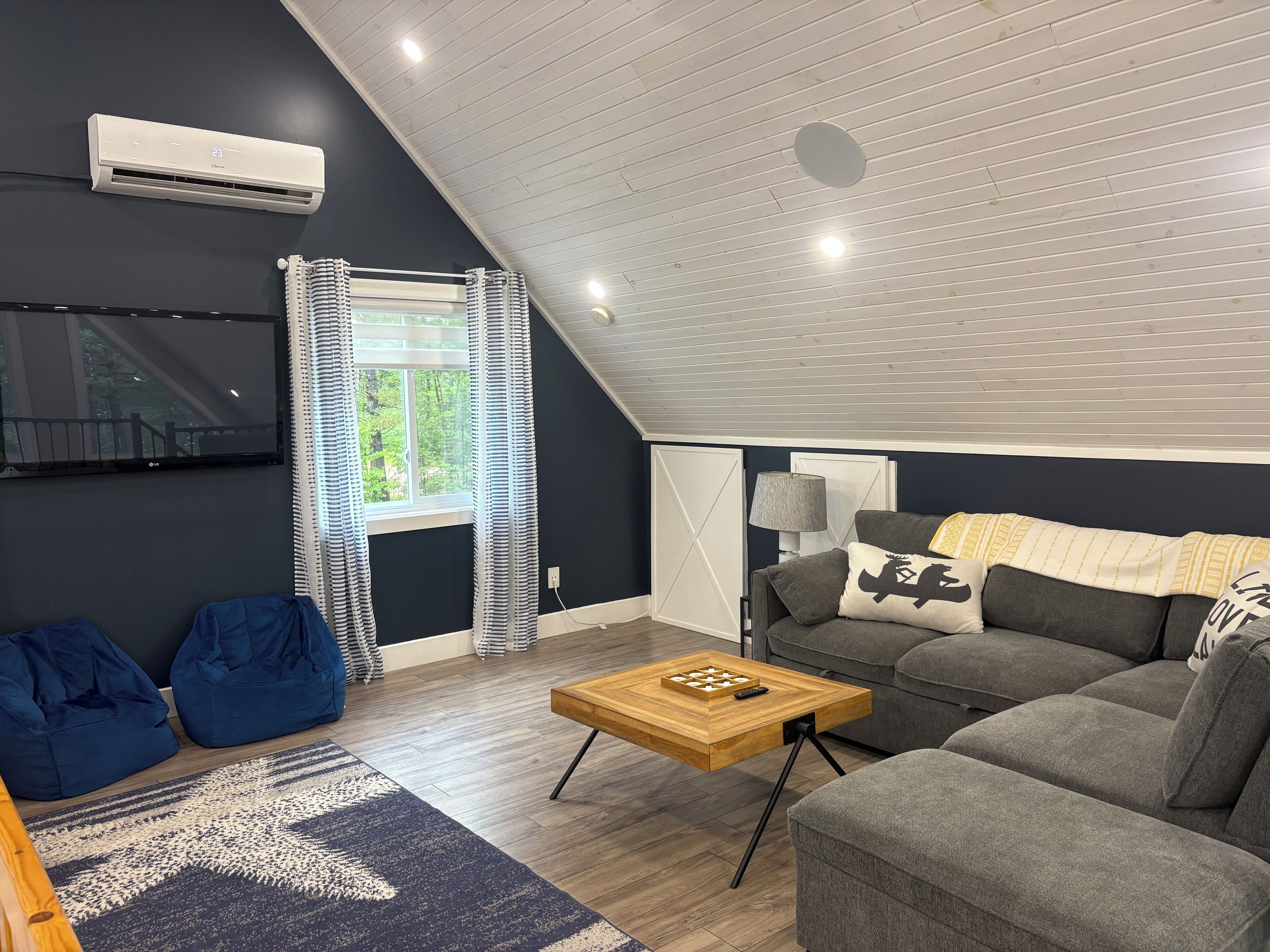 A modern living room with a sloped ceiling, a large window with curtains, a grey sofa, blue bean bag chairs, a wooden coffee table, and a TV mounted on the wall.