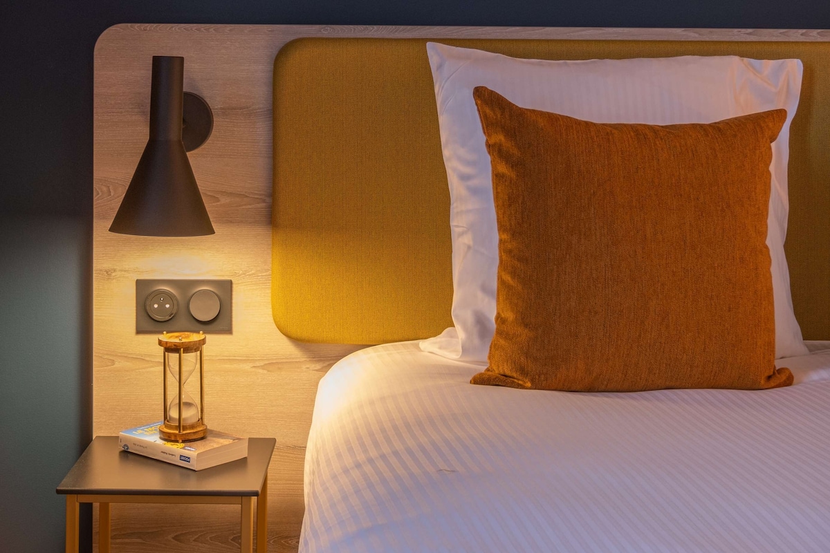 Een net opgemaakt bed met een wit en bruin kussen, een houten nachtkastje met een lamp, en een bedieningspaneel aan de muur.