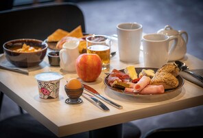 A breakfast setting with a plate of meats, cheese, and bread, a bowl of soup, a cup of coffee, and a glass of beer on a wooden table.