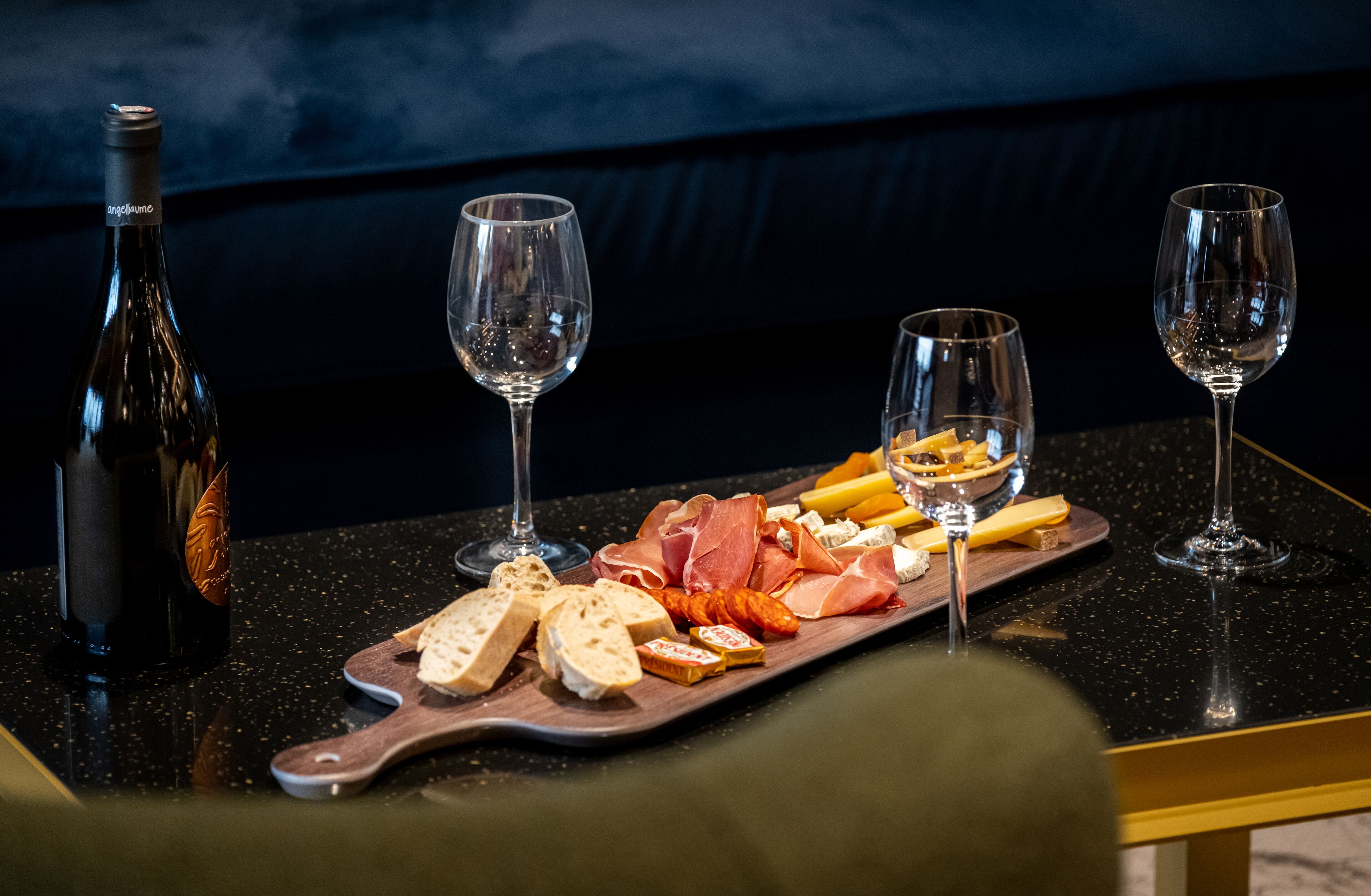 Une table avec une bouteille de vin, deux verres et une planche en bois sur laquelle sont disposés différents fromages et différentes charcuteries.