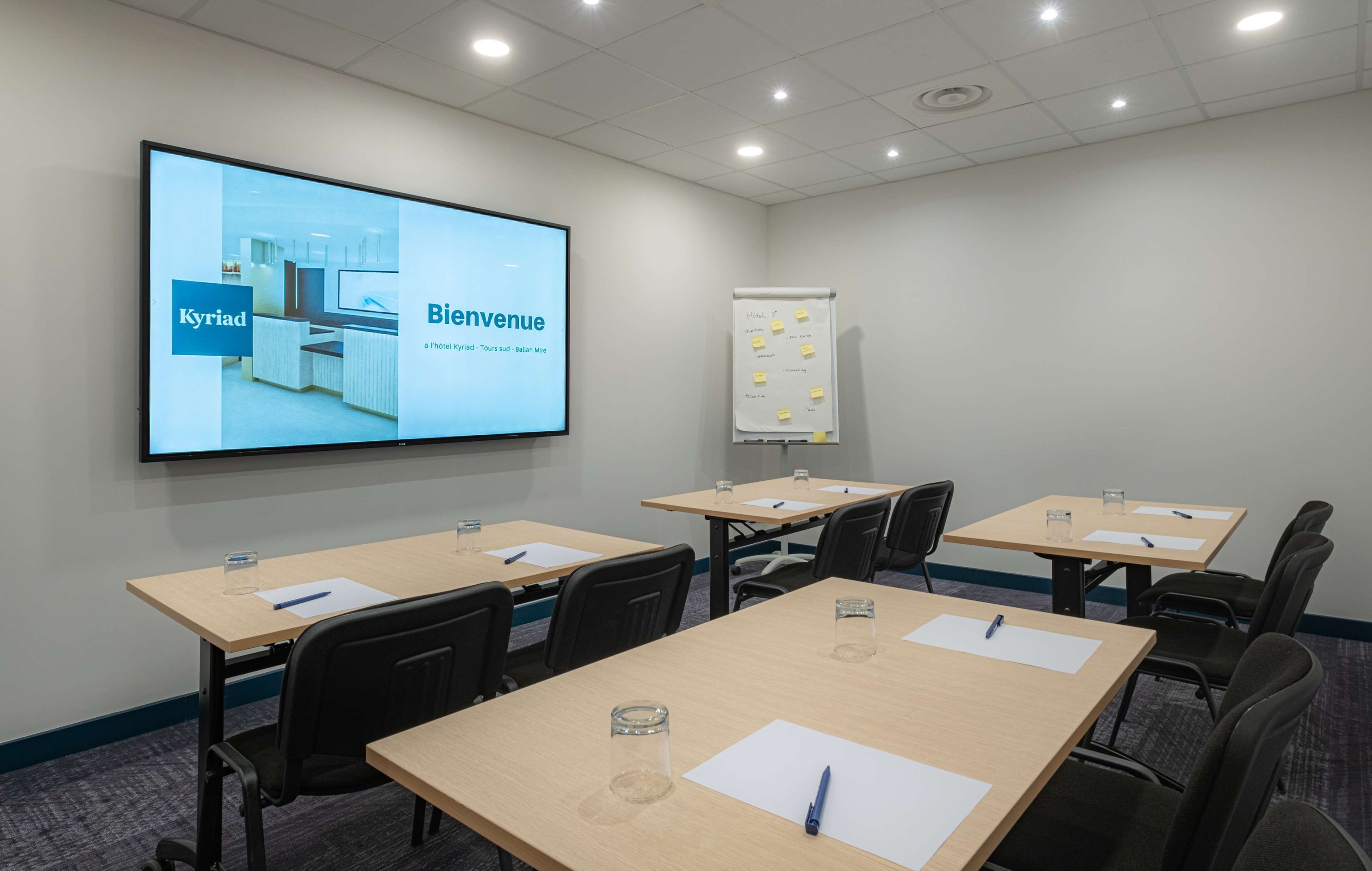 Un salón de conferencias con una pantalla grande mostrando el interior de un hotel, una pizarra con notas adhesivas y mesas con bolígrafos y blocs de notas.