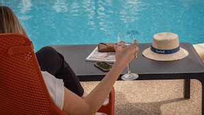 Una persona descansando junto a una mesa en la piscina, con una bebida, un sombrero y una revista.