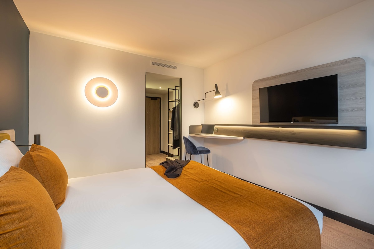 Een moderne hotelkamer met een groot bed, een flatscreen televisie aan de muur, een bureau met een laptop en een ronde lamp.