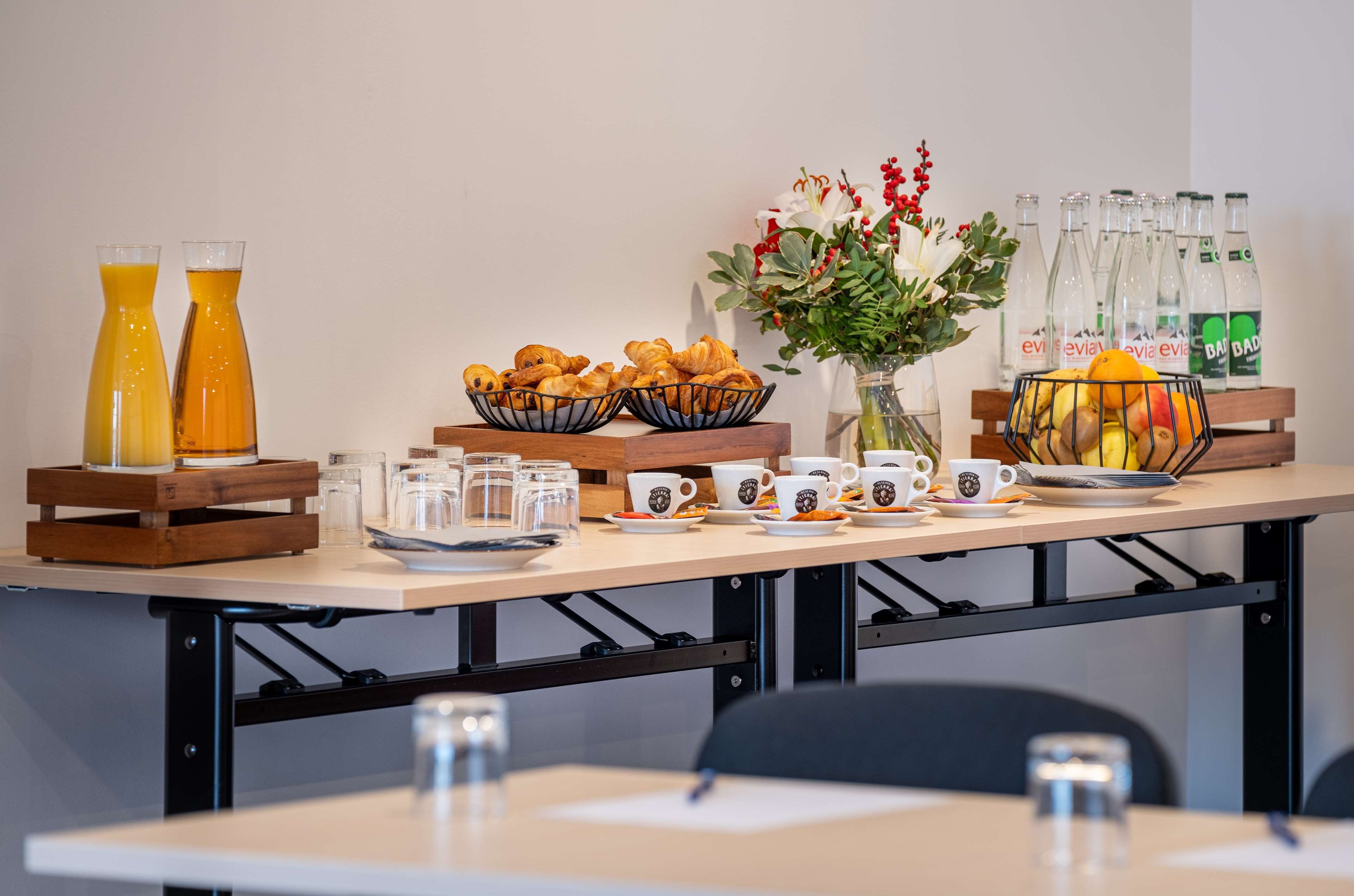 A breakfast setup with croissants, juice, coffee cups, and a fruit bowl on a table.