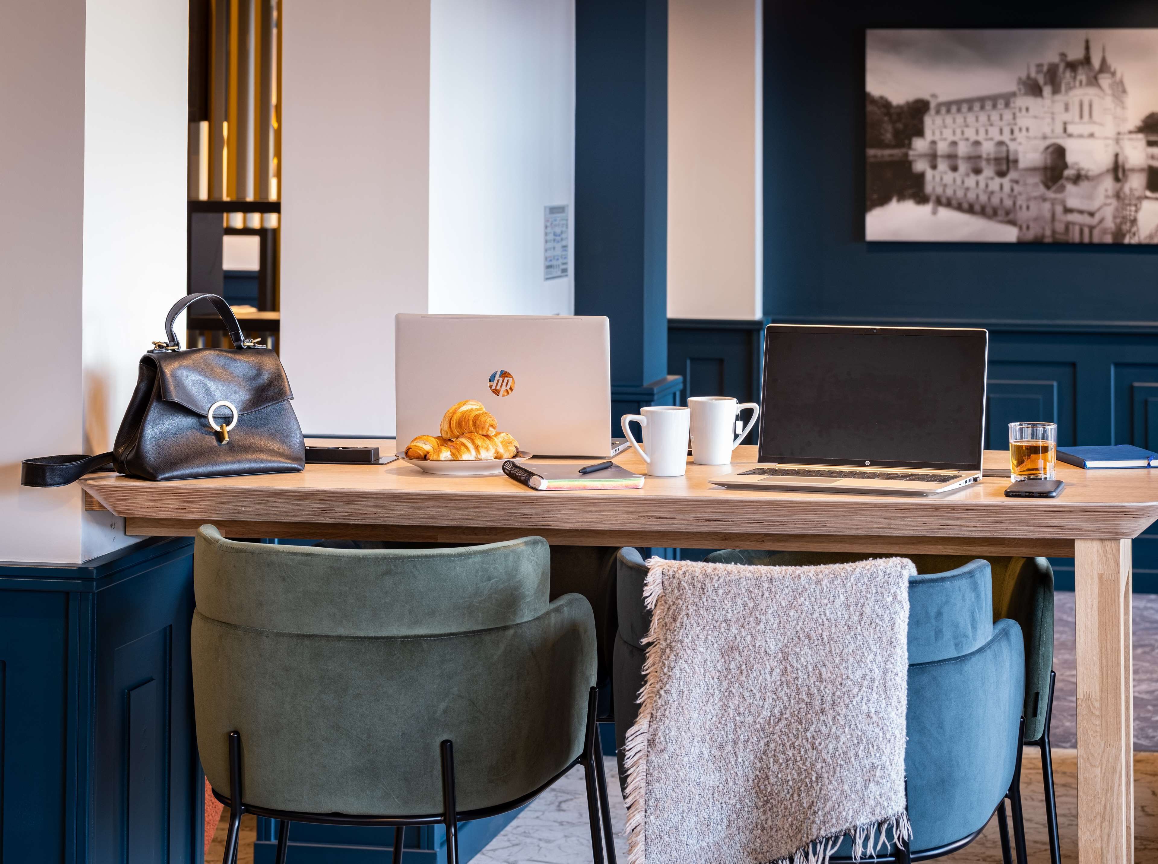 A wooden desk with laptops, a handbag, croissants, and coffee cups.