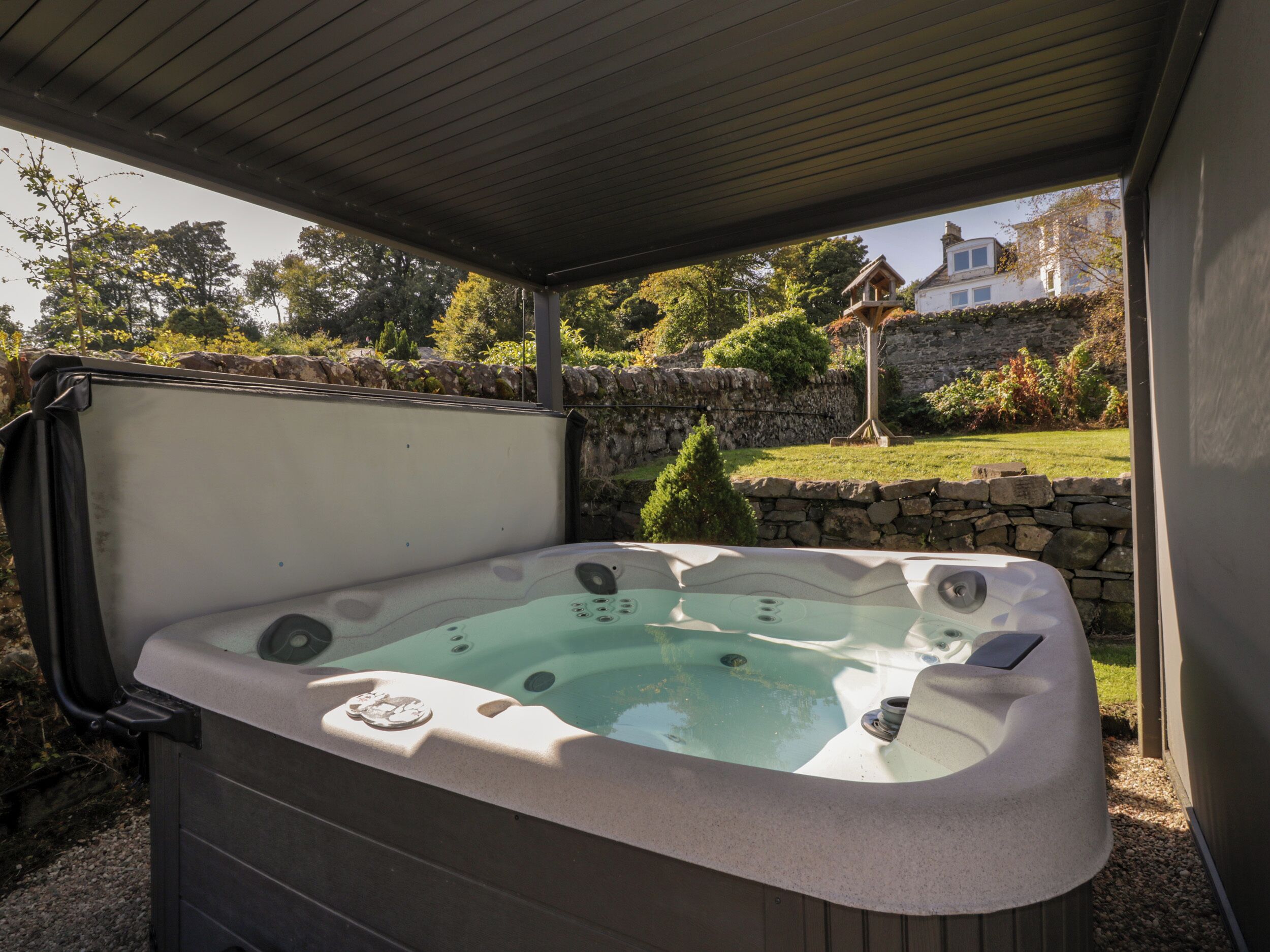 Un jacuzzi bajo un porche cubierto, con una pared de piedra y follaje al fondo.