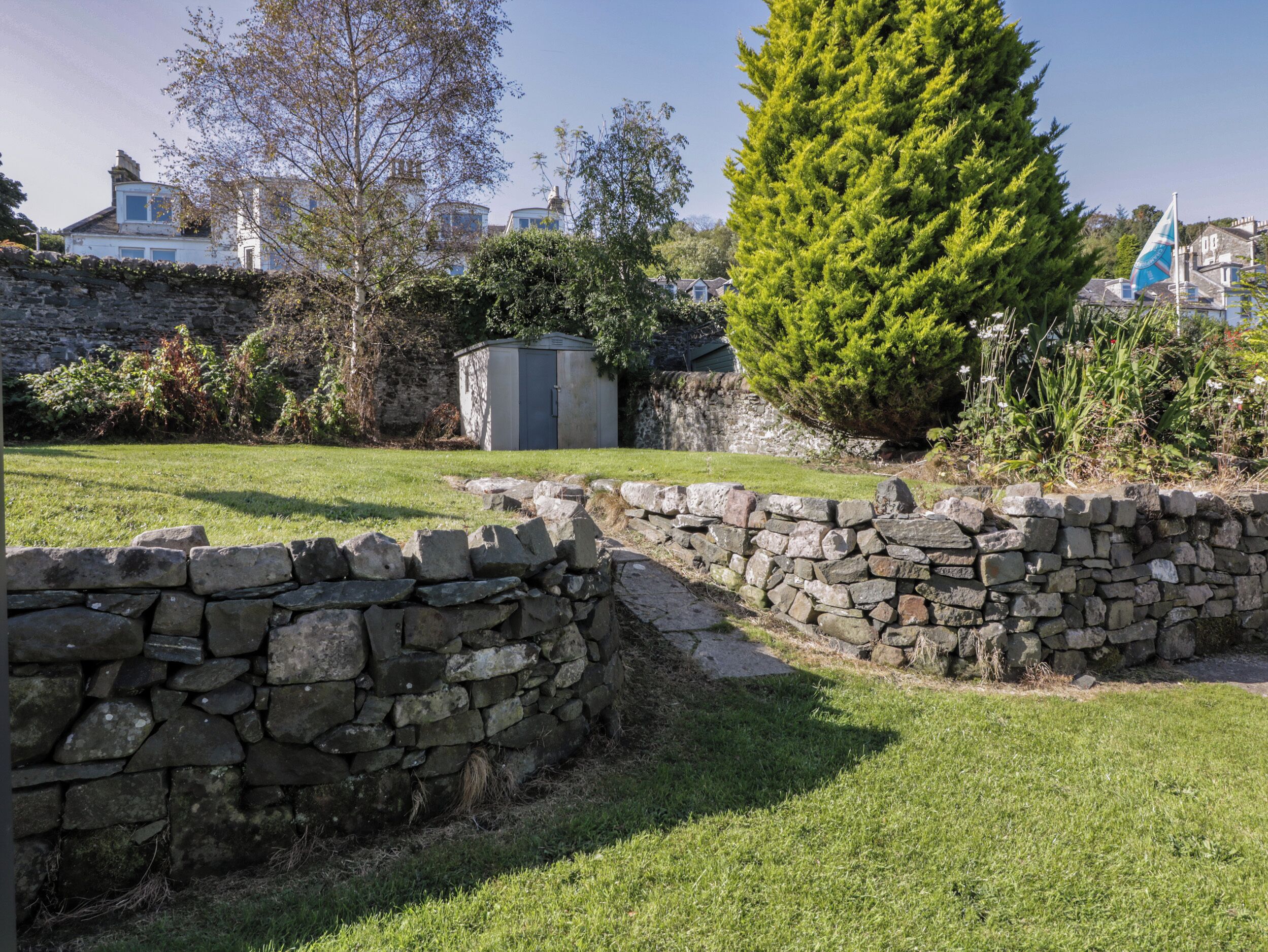 Un mur de pedra, una petita caseta i un arbre conífer alt en un jardí.
