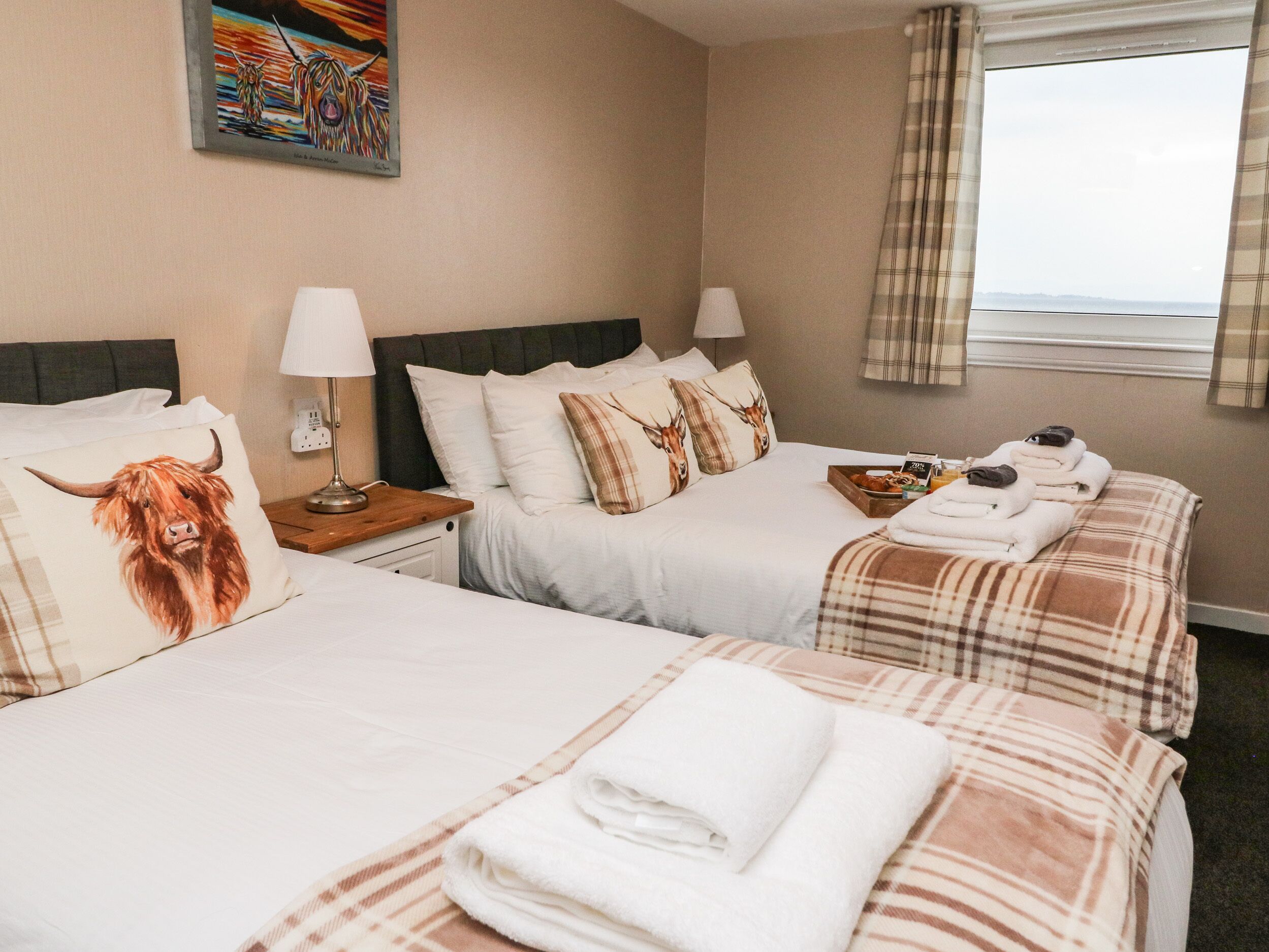 A hotel room with two beds, each with a pillow featuring a bull's head, a bedside table with a lamp, a window with curtains, and a view of the sea.