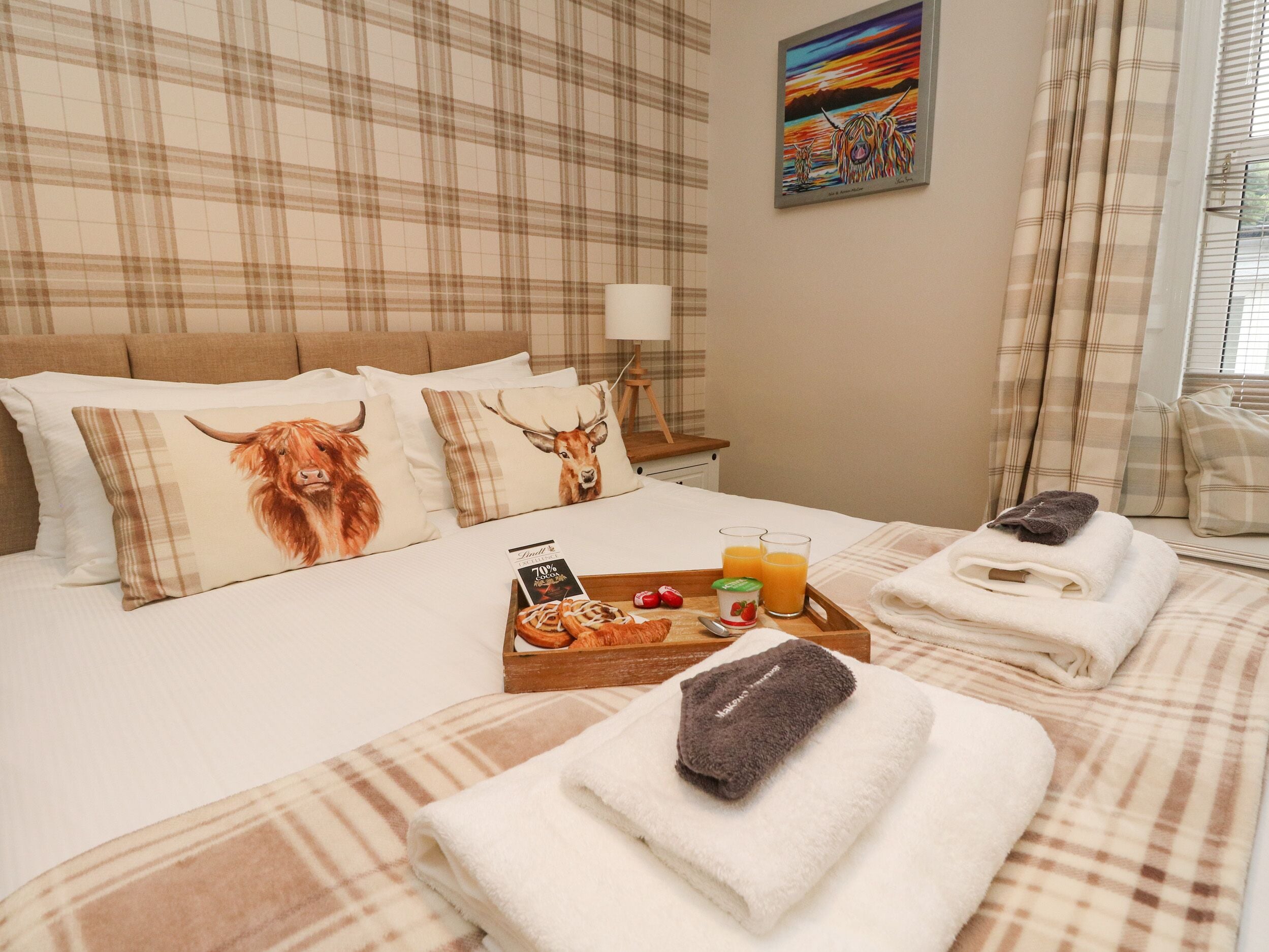 Camera d'albergo con un letto con biancheria a quadri, due cuscini a fantasia animalier, un vassoio con prodotti per la colazione e un quadro appeso al muro.