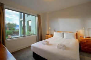 Une chambre d’hôtel avec une grande fenêtre, un lit avec du linge de lit blanc, deux tables de chevet et une petite table de nuit.