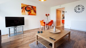 Ett modernt vardagsrum med en platt-TV, ett soffbord i trä, orangefärgade stolar och ett matbord. En stor, abstrakt målning och en väggklocka syns.