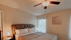Un dormitorio con tablero de cabecera capitonado, cama con colcha a rayas, mesita de noche con lámpara, ventilador de techo y ventana con cortinas traslúcidas.