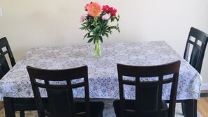 Una mesa de comedor con mantel floral, rodeada por cuatro sillas de madera. Un jarrón con flores está sobre la mesa.