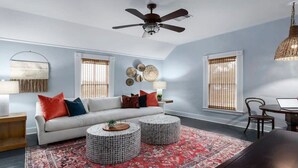 Un salon avec un canapé blanc, deux petites tables rondes, un tapis rouge à motifs et un ventilateur de plafond.