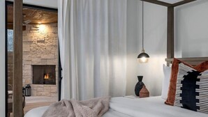 Une chambre avec une cheminée en pierre, un lit recouvert d’une couverture rose et une table de chevet avec une lampe.