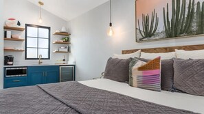 Une chambre moderne avec un lit, des tables de chevet, une fenêtre et un coin cuisine doté de meubles bleus et d’un évier.