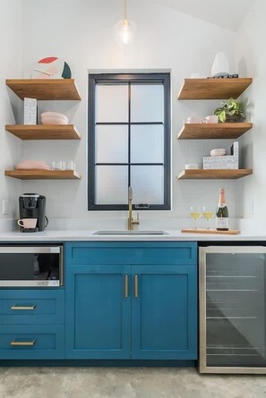 A kitchen with teal cabinets, a window, wooden shelves, and a coffee maker.