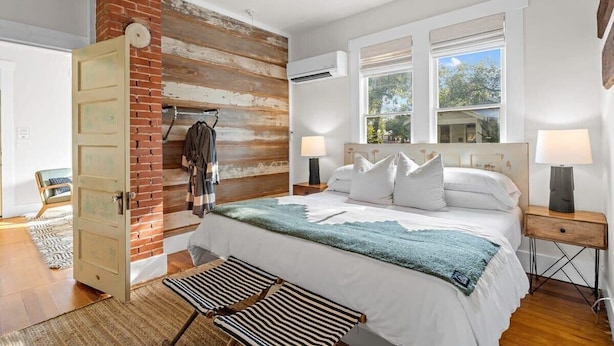 Ein Schlafzimmer mit Backsteinwand, einem Bett mit weißer Bettwäsche, zwei Nachttischlampen und einem Fenster mit Blick auf Bäume.