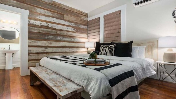 Une chambre avec un mur en bois, un lit recouvert d’une couverture à rayures noires et blanches, une table de chevet avec une lampe et un banc.