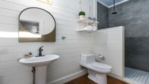 Une salle de bain avec un lavabo blanc, des toilettes et un espace douche. On y trouve des étagères avec des serviettes et une petite plante.