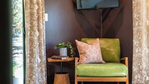 Ein Zimmer mit einem grünen Ledersessel, einem kleinen Holznachttisch mit einem Buch und einer Pflanze sowie einem an der Wand montierten Fernseher.