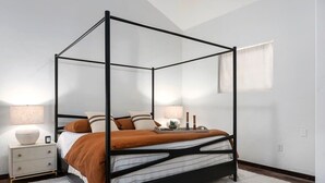 A four-poster bed with a white and brown color scheme, a bedside table with a lamp, and a small plant on the nightstand.