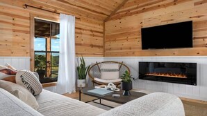 A cozy cabin interior with a sofa, a wicker chair, a fireplace, and a TV mounted on the wall.