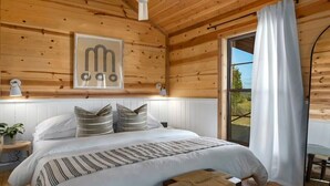 A wooden cabin bedroom with a bed, pillows, a framed picture, and a window with curtains.