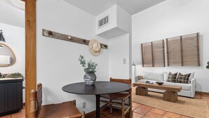 A dining area with a round table, wooden chairs, and a vase with plants. There is a sofa and a window with blinds in the background.