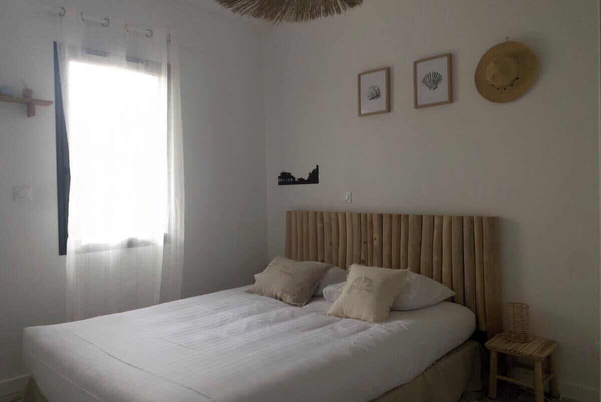 A bedroom with a bed, a wooden headboard, a small wooden stool, and a window with sheer curtains.