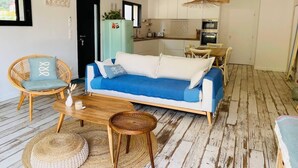 Un salon moderne avec un canapé bleu, des fauteuils en osier, une table basse en bois et une cuisine en arrière-plan.
