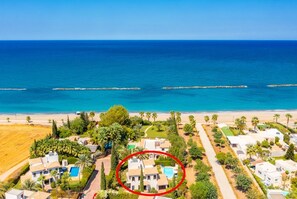 Una zona residencial costera con una amplia playa, palmeras y casas con piscinas.