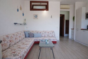 A living room with a sofa, a small table, and a kitchen area with cabinets and a microwave. - Scirocco House | Terrace with a view of the sea (Isola Rossa)