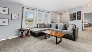 A living room with a sectional sofa, a coffee table, and a small wooden side table. There are framed pictures on the wall and a window with blinds. - Located near Mt. Rushmore, the Black Hills, Badlands National Park & more!! (Hermosa)